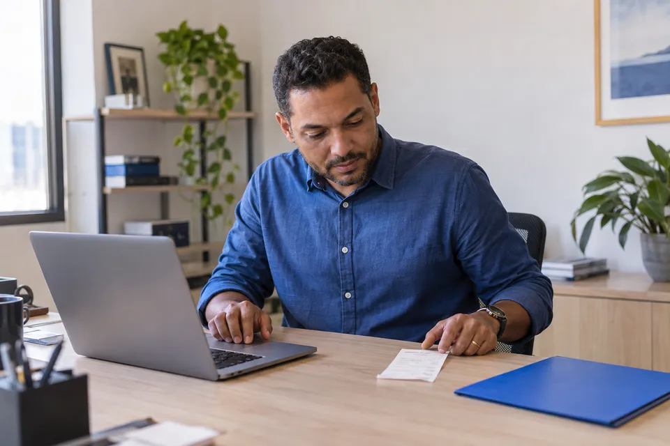 Gestor conferindo um comprovante ao lado do notebook no escritório.